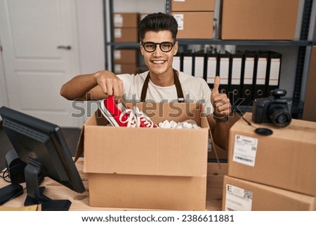 Young hispanic man working at small business ecommerce smiling happy and positive, thumb up doing excellent and approval sign 