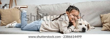 happy girl smiling and lying on sofa with cute labrador in modern living room, pet and kid Royalty-Free Stock Photo #2386614499