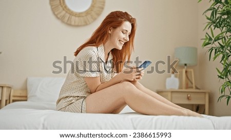 Young redhead woman using smartphone sitting on bed at bedroom