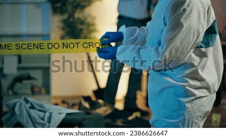 A forensic expert is marking the murder scene with warning tape, and police detectives are investigating the crime Royalty-Free Stock Photo #2386626647