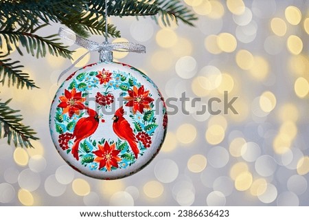 Christmas memorial ornament red cardinal hanging from a tree with golden lighting with bokeh in the background. Ukrainian folk petrykivka art.