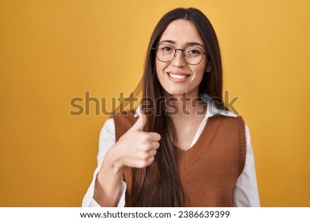 Young brunette woman standing over yellow background wearing glasses doing happy thumbs up gesture with hand. approving expression looking at the camera showing success. 