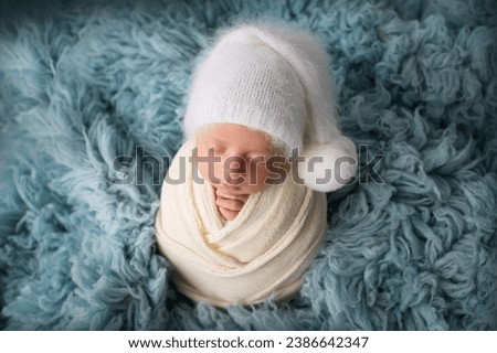 Sleeping newborn baby boy in the first days of life in a white soft cocoon with a knitted woolen white hat on a blue background. Studio macro photography, portrait of a newborn. Woman's happiness. 