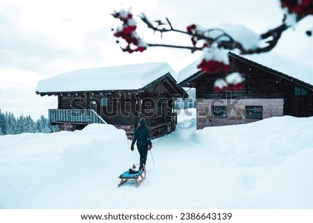 A Caucasian adult male is pictured pulling a wooden toboggan sled outside of a small log cabin in a winter landscape