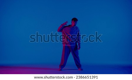 A young man stands against a blue background and looks at the camera. A pink light shines on him from the side and illuminates one side. He is a dancer performing a dance and showing the moves