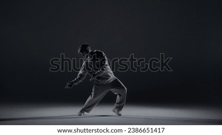 A young man stands against a dark background. He is highlighted by a white light from below. Creates a light haze. Demonstrates a dance movement by leaning to the side. He is plastic, rhythmic