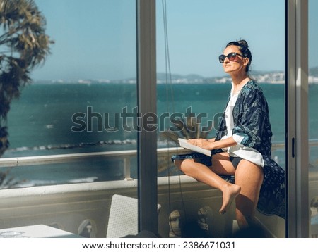 Through doorway side view of optimistic female reading book on balcony of hotel room against sea during summer trip in tropical country Royalty-Free Stock Photo #2386671031
