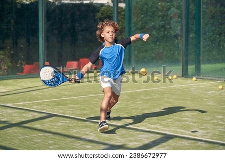 Full body of active boy in activewear hitting ball with racket while playing padel game on sports ground on summer day Royalty-Free Stock Photo #2386672977