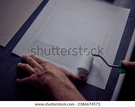 Crop artisan holding roller and plastering sheet of marked paper with professional bookbinding glue