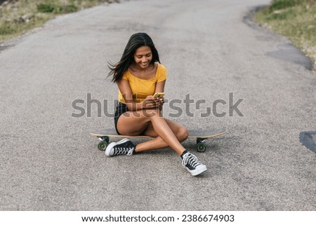 Full body of positive ethnic female browsing on smartphone while sitting longboard on asphalt road