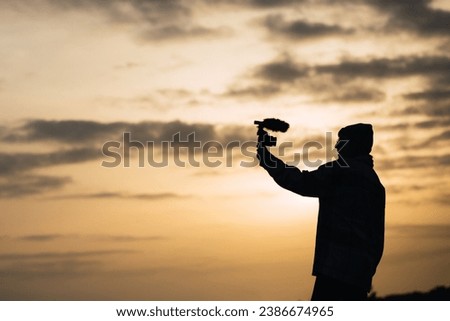 Side view silhouette of unrecognizable young female blogger in hat shooting video on digital camera against sunset sky in countryside