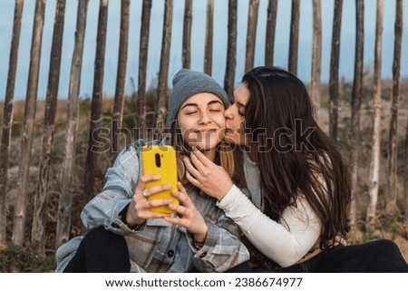 Young lady with long dark hair in casual clothes sitting on wooden path in rural terrain and hugging and kissing cheek of female best friend using smartphone