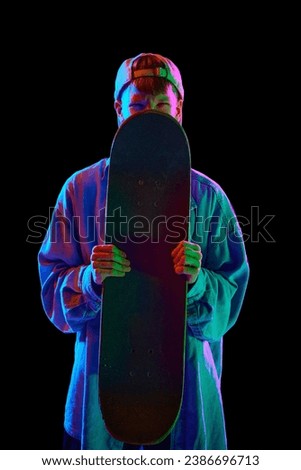 Teen boy in casual clothes standing with skateboard, posing against black studio background in neon light. Concept of professional sport, competition, training, action. Copy space for ad