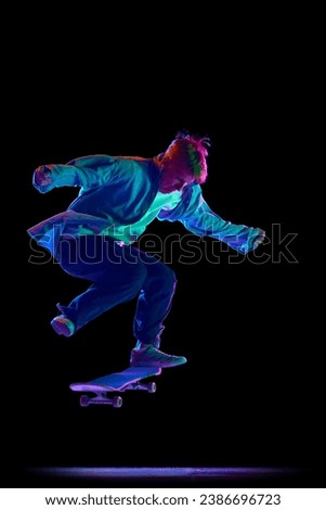 Teen boy in blue jeans shirt and cap in motion, training with skateboard, doing stunts against black studio background in neon light. Concept of professional sport, competition, training, action.