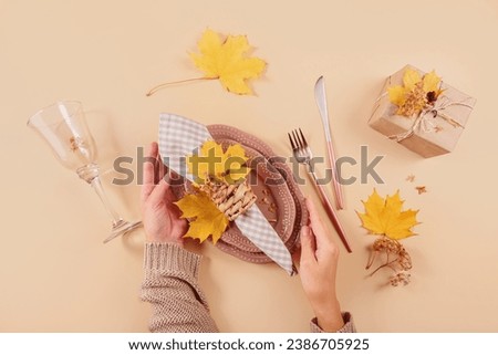 Autumn table setting on beige background. Womens hands holding oval plates, cutlery, checkered napkin, gift box and dry yellow leaves. Autumn holiday mood, Halloween, Thanksgiving concept. Top view