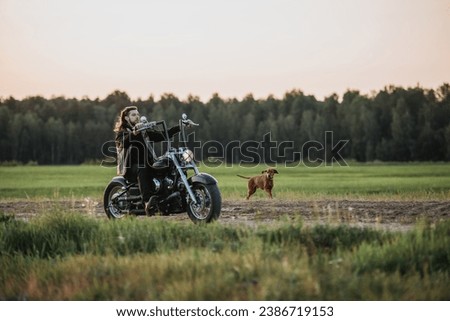 Young handsome long-haired man riding on motorbike at countryside road with his dog runs nearby Royalty-Free Stock Photo #2386719153
