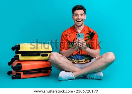 Full size photo of impressed crazy guy wear tropical shirt stylish shorts hold smartphone sit near bags isolated on blue color background
