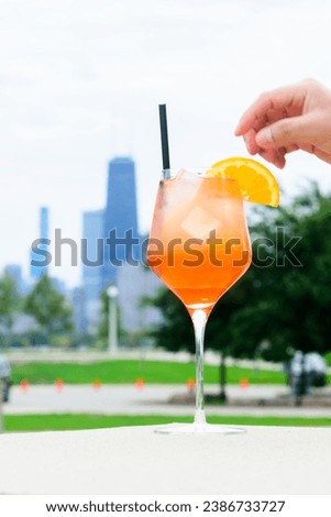 A hand garnishes an orange cocktail with a slice in a stemmed glass against a city skyline Royalty-Free Stock Photo #2386733727