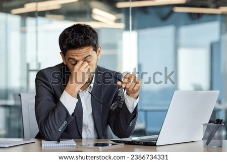 Overtired and overwhelmed businessman at workplace inside office, man took off glasses rubbing eyes, dizziness migraine and headache, man in business suit working late with laptop at workplace. Royalty-Free Stock Photo #2386734331