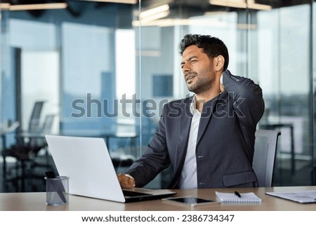 Neck pain, overworked and overtired man working late, businessman massaging neck muscle, working sitting on chair at table with laptop inside office at workplace. Royalty-Free Stock Photo #2386734347