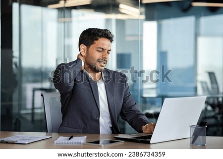 Neck pain, overworked and overtired man working late, businessman massaging neck muscle, working sitting on chair at table with laptop inside office at workplace. Royalty-Free Stock Photo #2386734359
