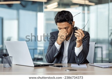 Overtired and overwhelmed businessman at workplace inside office, man took off glasses rubbing eyes, dizziness migraine and headache, man in business suit working late with laptop at workplace. Royalty-Free Stock Photo #2386734373