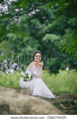 portrait of a happy woman or bride sitting under a shady tree, with makeup and wearing a white wedding dress. In the photo outdoors with trees in the background.