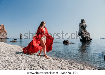 Woman travel sea. Happy tourist in red dress enjoy taking picture outdoors for memories. Woman traveler posing on the rock at sea bay surrounded by volcanic mountains, sharing travel adventure journey