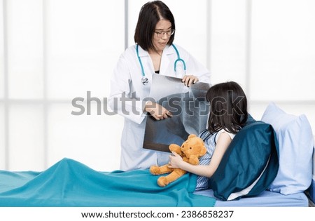 Asian professional friendly female doctor in white lab coat and stethoscope standing smiling showing lungs Xray film to young girl patient lay down on bed hugging teddy bear doll in hospital wardroom.