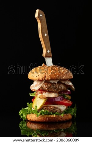 Fresh tasty burger with knife on a black reflective background. Burger with tomato, onion, preserved cucumber, salad, cheese, beef cutlet, and bacon.