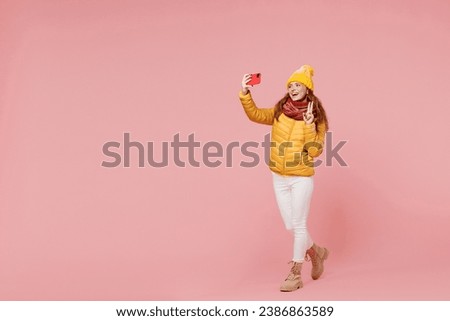 Full size body length fascinating young woman 20s wears yellow jacket hat mittens do selfie shot on mobile cell phone show victory sign isolated on plain pastel light pink background studio portrait