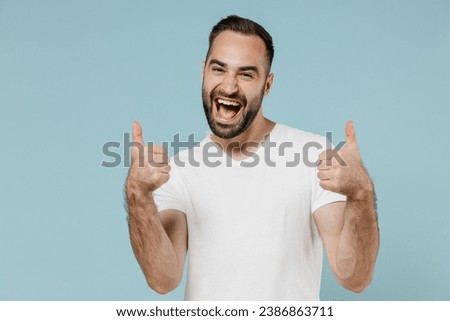 Young exultant happy satisfied man 20s in blank print design casual white t-shirt show thumb up gesture isolated on plain pastel light blue color background studio portrait. People lifestyle concept