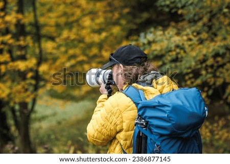 Hiker with backpack is wearing yellow jacket and photographing at his camera. Nature or landscape photographer in autumn forest