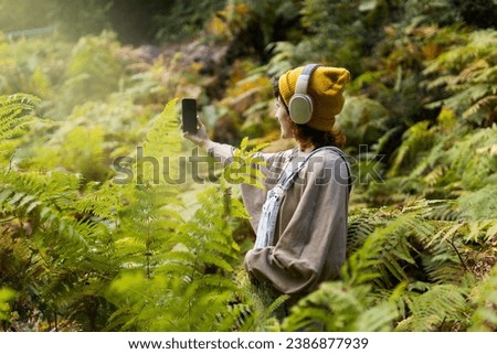 Woman with mobile phone and headphones in fern forest wearing yellow hat, listening to music, taking photos and video call