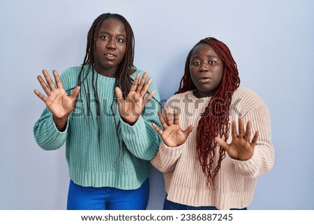 Two african woman standing over blue background moving away hands palms showing refusal and denial with afraid and disgusting expression. stop and forbidden. 