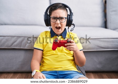 Young hispanic kid playing video game holding controller wearing headphones angry and mad screaming frustrated and furious, shouting with anger. rage and aggressive concept.  Royalty-Free Stock Photo #2386889029