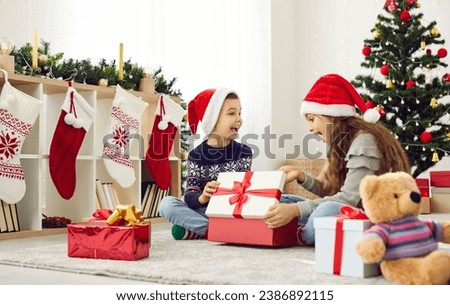 Overjoyed kids in Santa hats feel excited open Christmas presents on winter holiday morning at home. Smiling children near fir-tree have fun unpack unbox gifts on New Year celebration together.