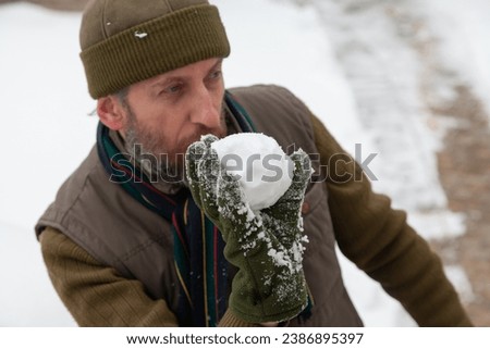 Snow games in the air. A man with a beard in a warm vest and a hat throws snowballs in the yard of a private house on a frosty day

