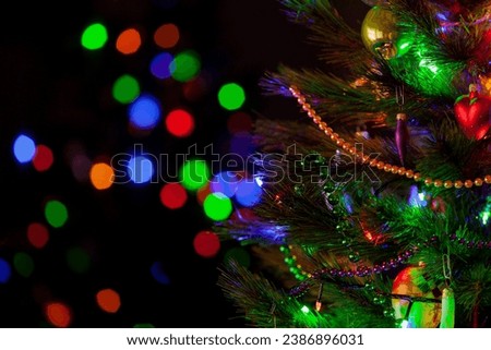 A green Christmas tree festively decorated with toys and luminous garlands stands in a dark room close-up. In the background, colorful bokeh lights



