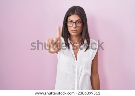 Brunette young woman standing over pink background wearing glasses pointing with finger up and angry expression, showing no gesture 