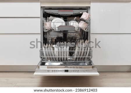 Open built-in dishwasher machine with clean cutlery, dishes, plates in white modern kitchen. front view Royalty-Free Stock Photo #2386912403