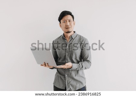 Asian man with beard wear grey shirt, dislike unsatisfied face, holding notebook hand gesture, unhappy question face looking at camera isolated over white background wall. Royalty-Free Stock Photo #2386916825