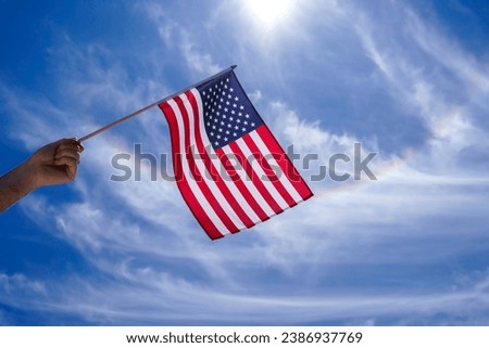 Person waving an American flag with a rainbow color streak of light and clouds in the background. The United States, USA flag is part of the American culture. It is often flown on the July 4th holiday