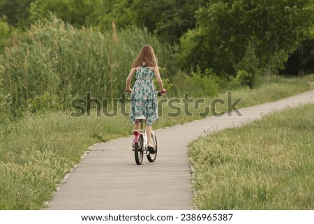A girl rides a bicycle on an asphalt avenue among the greenery of the park. Active fun outdoors. Sports load for the proper development of the girl's body.