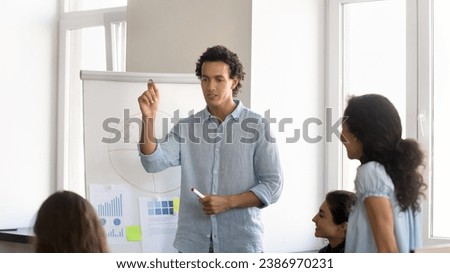 Serious handsome young employee sharing sales report data with colleagues, presenting statistic graphs at whiteboard. Mentor, coach, project leader man speaking to intern on office meeting