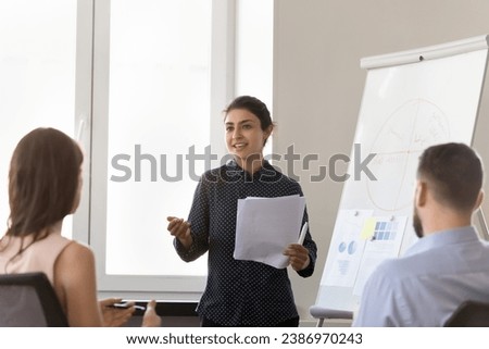 Happy motivated Indian corporate teacher woman training interns, teaching employees, coaching team of students, speaking at marketing charts on white board on office meeting Royalty-Free Stock Photo #2386970243