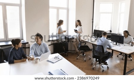 Multiethnic business team working in small office space, using laptop computer, cooperating on project, common tasks, talking at shared workplace tables. Modern rental co-working space