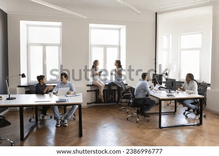 Diverse busy employees working on modern office room, sitting at workplaces, using desktops, talking, discussing project tasks. Contemporary rental office co-working space Royalty-Free Stock Photo #2386996777