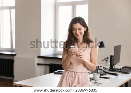Cheerful happy pretty mobile phone user woman chatting online, using app on cellphone for Internet communication, reading text message, typing, standing at office workplace table, smiling, laughing