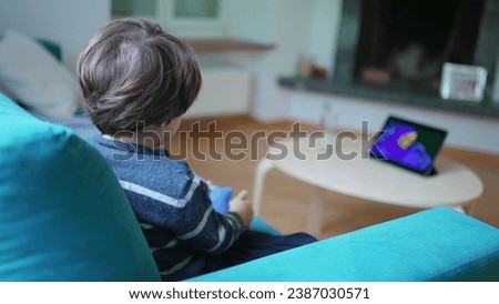 Child watching cartoon at home seated on sofa couch, small boy absorbed by entertainment content on tablet device screen